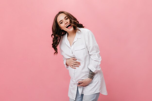Retrato de mujer feliz emocional en camisa larga blanca. Señora embarazada alegre en jeans riendo y posando sobre fondo rosa.