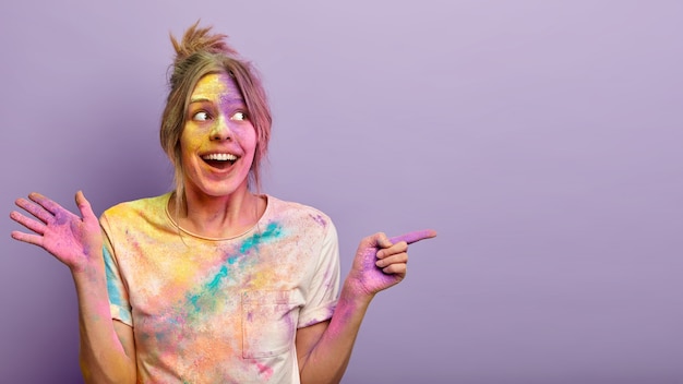 Retrato de mujer feliz y encantada con la cara sucia y ropa con tintes de colores, celebra el festival de Holi en la India, demuestra el espacio libre sobre una pared púrpura. Fiesta de colores. Concepto de promoción