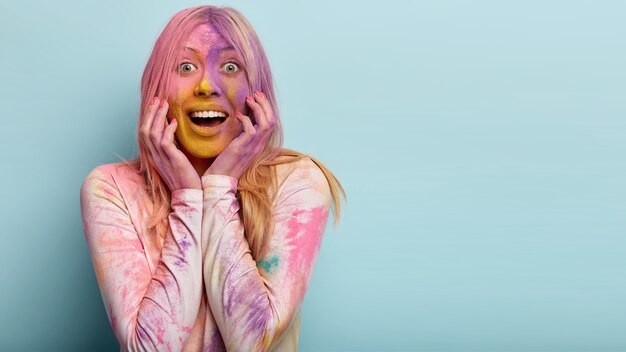 Retrato de mujer feliz y encantada sonríe positivamente, muestra dientes blancos, tiene expresión amistosa, feliz de estar en el festival Holi Colors, empolvado con tinte de colores, se encuentra en el interior, espacio en blanco para texto