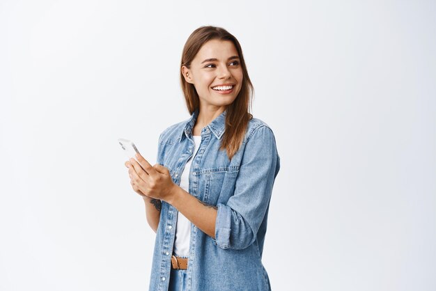Retrato de una mujer feliz y guapa con cabello rubio mira directamente al espacio de la copia sosteniendo un teléfono inteligente chateando en las redes sociales o usando una aplicación móvil de fondo blanco
