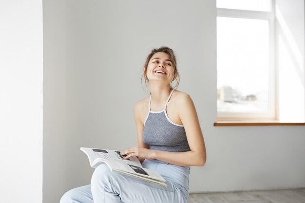 Retrato de la mujer feliz hermosa joven que sonríe riendo sosteniendo el libro que se sienta en silla sobre la pared blanca en casa.