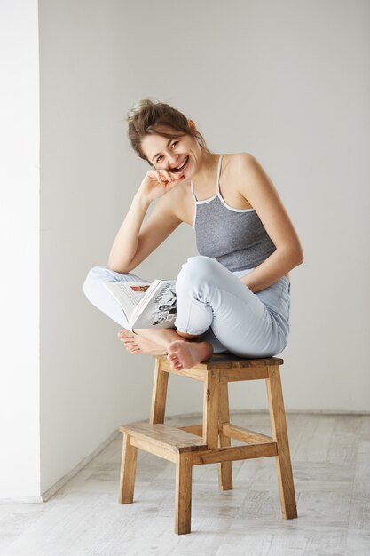 Retrato de la mujer feliz hermosa joven que sonríe riendo sosteniendo el libro que se sienta en silla sobre la pared blanca en casa.