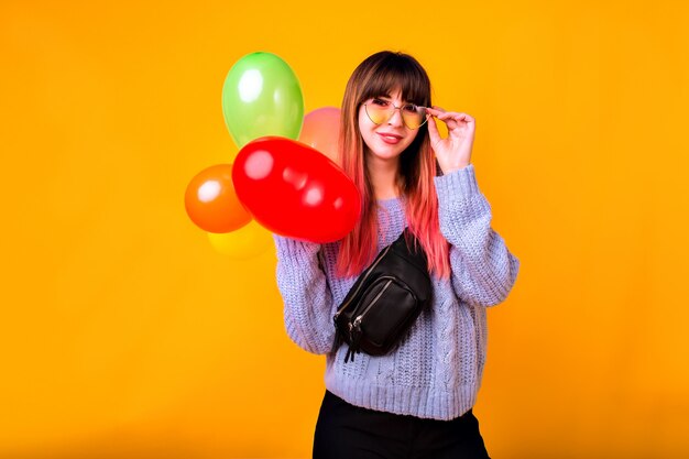 Retrato de mujer feliz joven inconformista mostrando gesto ok y riendo, suéter azul acogedor, gafas de moda y bolso, sosteniendo globos de colores, humor de fiesta.