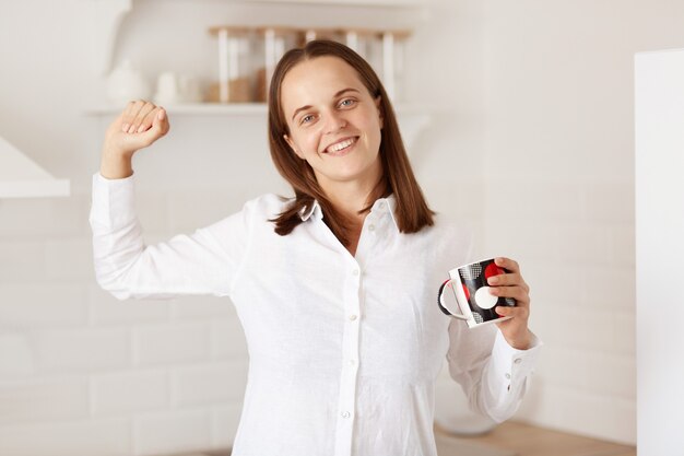 Retrato de mujer feliz de pie con el brazo levantado, sosteniendo una taza con té, estirando las manos después de despertarse, mirando a la cámara con una sonrisa encantadora, vistiendo una camisa blanca de estilo casual.