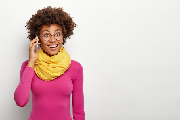 Retrato de mujer feliz de piel oscura con peinado afro, usa anteojos, poloneck y bufanda amarilla alrededor del cuello, tiene expresión de rostro alegre, modelos sobre la pared blanca del estudio