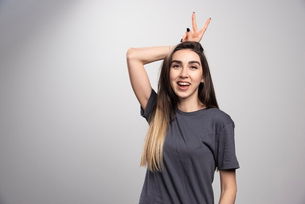 Retrato de mujer feliz posando y poniendo orejas de conejo sobre fondo gris.