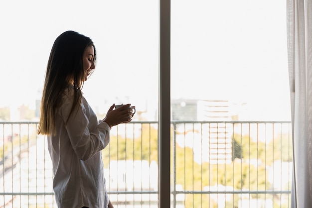 Retrato de una mujer feliz que se coloca en el balcón que sostiene la taza de café disponible