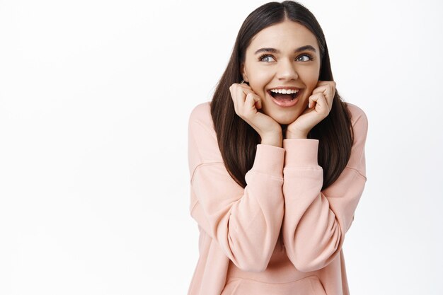 Retrato de mujer feliz soñadora imaginando algo, apoyado en las manos y sonriendo pensativo, mirando la esquina superior izquierda con interés, soñando despierto, pared blanca