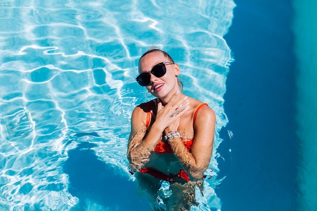 Retrato de mujer feliz tranquila en gafas de sol con piel bronceada en piscina azul en día soleado