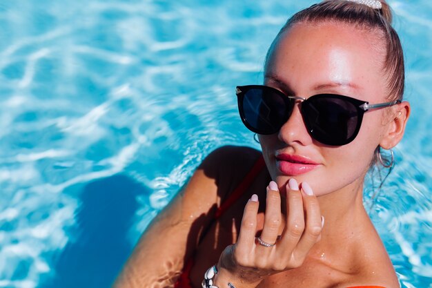 Retrato de mujer feliz tranquila en gafas de sol con piel bronceada en piscina azul en día soleado