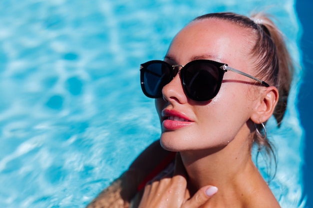 Retrato de mujer feliz tranquila en gafas de sol con piel bronceada en piscina azul en día soleado