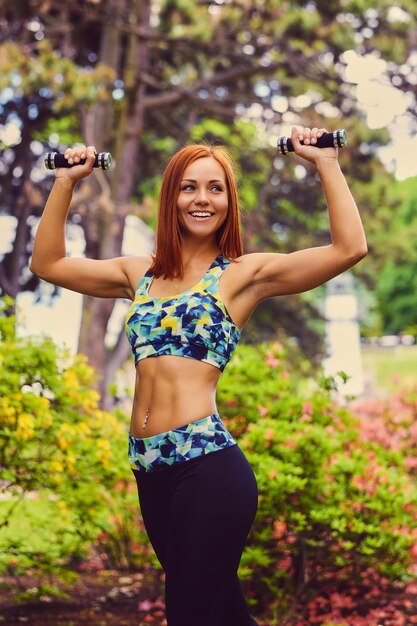 Retrato de mujer fitness pelirroja sostiene pesas en un parque al aire libre.