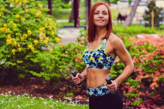 Retrato de mujer fitness pelirroja sostiene pesas en un parque al aire libre.