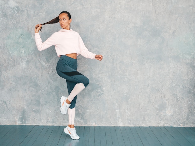 Retrato de mujer de fitness en ropa deportiva que parece segura. Joven mujer vistiendo ropa deportiva. Hermosa modelo con cuerpo bronceado perfecto. Salto femenino en estudio cerca de la pared gris.