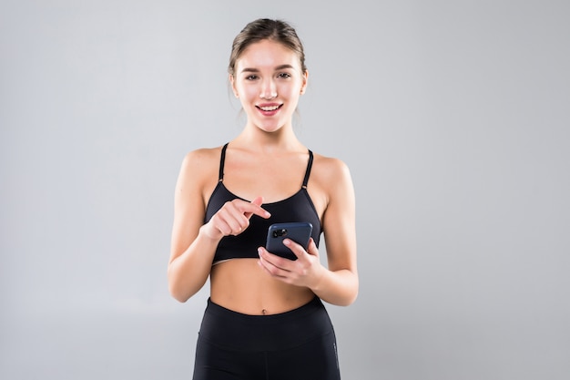 Retrato de una mujer fitness sonriente enviando mensajes de texto en el teléfono móvil y con auriculares en una pared blanca