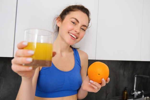 Foto gratuita retrato de una mujer de fitness sonriente ofreciendo jugo de naranja con fruta y un vaso en las manos posando en