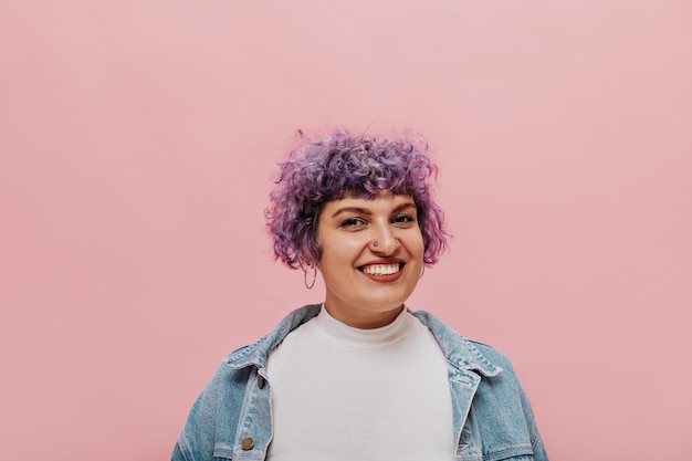 Foto gratuita retrato de mujer fresca con pelo morado corto y rizado con piercing y sonrisa en rosa aislado.