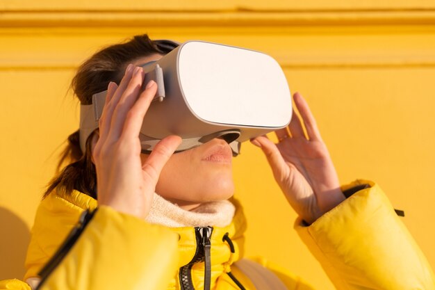 Retrato de una mujer con gafas de realidad virtual en la calle contra una pared amarilla a la luz del sol en invierno en ropa de abrigo