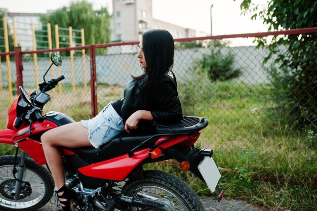 Retrato de una mujer genial e impresionante con vestido y chaqueta de cuero negro sentada en una moto roja genial