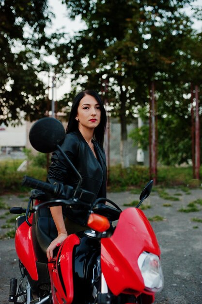 Retrato de una mujer genial e impresionante con vestido y chaqueta de cuero negro sentada en una moto roja genial