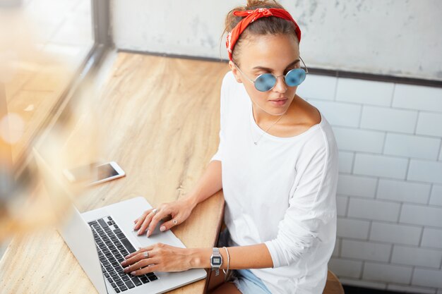 Foto gratuita retrato de mujer gerente trabaja en plan de negocios en computadora portátil, busca información, mira pensativamente a un lado, rodeada de modernos aparatos electrónicos. mujer editora comprueba el contenido del sitio web