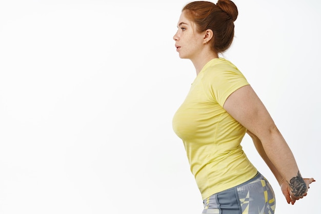 Foto gratuita retrato de mujer gordita pelirroja en ropa de fitness estirando los brazos detrás de la espalda haciendo ejercicio en el gimnasio de pie sobre fondo blanco.