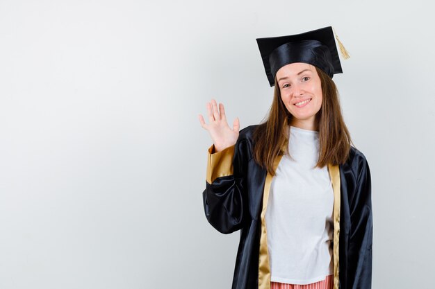 Retrato de mujer graduada agitando la mano para saludar en traje académico y mirando alegre vista frontal