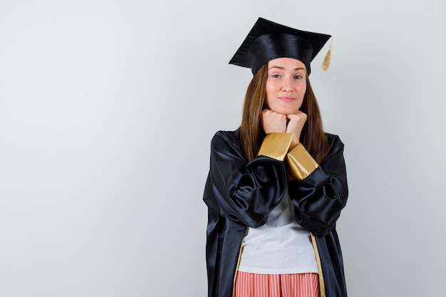 Retrato de mujer graduada apoyando la barbilla en los puños en uniforme, ropa casual y mirando inteligente vista frontal