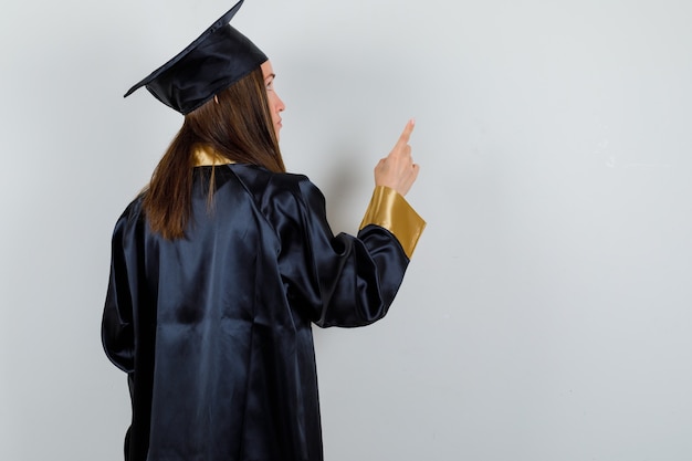 Retrato de mujer graduada apuntando hacia arriba en uniforme y mirando hacia atrás enfocada