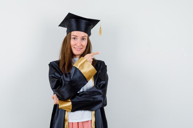 Retrato de mujer graduada apuntando a la derecha en traje académico y mirando esperanzada vista frontal