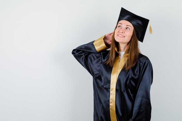 Retrato de mujer graduada posando mientras mira hacia arriba en traje académico y mira esperanzada vista frontal