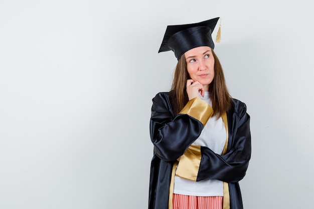Retrato de mujer graduada sosteniendo la mano en la barbilla en ropa casual, uniforme y mirando pensativo vista frontal
