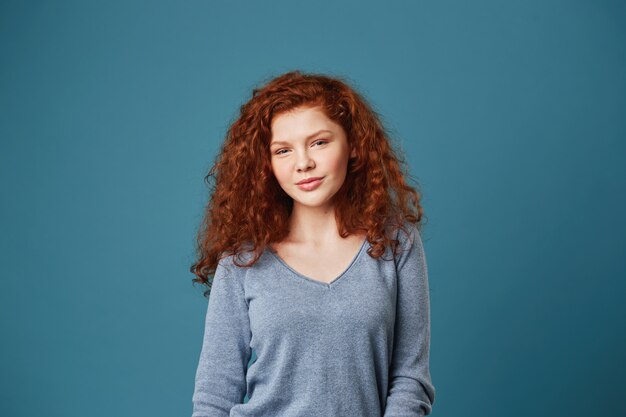 Retrato de mujer guapa estudiante con cabello rizado jengibre y pecas mirando con expresión tranquila y relajada.