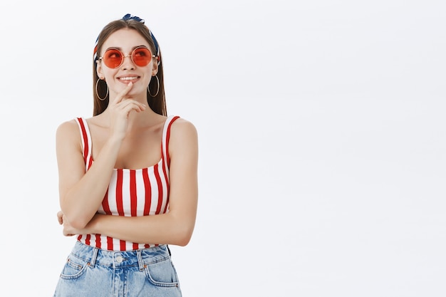 Retrato de mujer guapa feliz y pensativa creativa con gafas de sol y la parte superior a rayas tocando el labio mirando hacia arriba mientras toma una decisión contra la pared gris