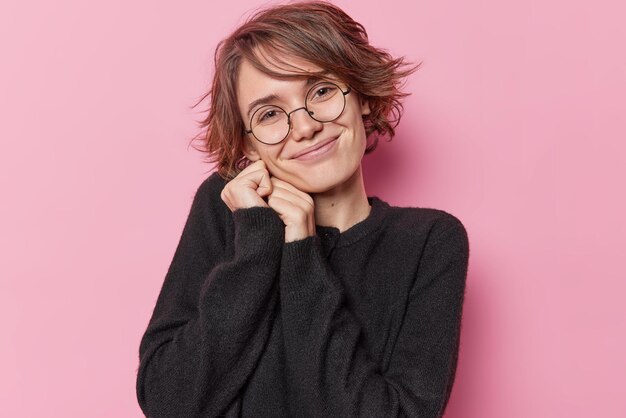 Retrato de una mujer guapa con un peinado moderno que se ve con admiración por algo que inclina la cabeza, mantiene las manos cerca de la cara, sonríe suavemente, usa anteojos redondos y un jersey negro aislado sobre una pared rosa