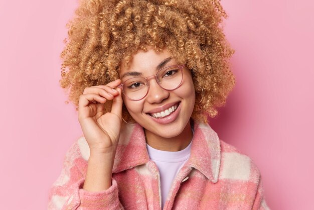 Retrato de una mujer guapa con el pelo rizado mantiene la mano en el borde de los espectáculos sonríe alegremente escucha noticias agradables vestida con una chaqueta informal aislada sobre un fondo rosa Concepto de emociones humanas