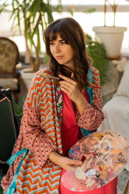 Retrato de mujer hermosa artista en traje bohemio posando con pincel y paleta en su estudio de arte.