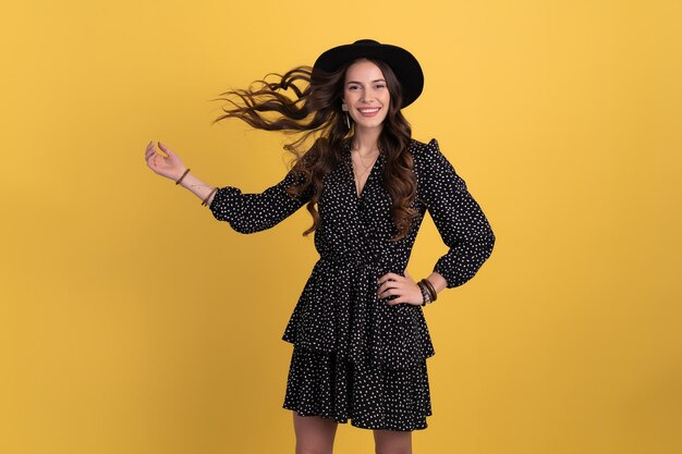 Retrato de una mujer hermosa y atractiva posando aislada en un fondo amarillo con un vestido negro punteado y un sombrero negro con estilo accesorios de estilo de moda de primavera verano sonriendo feliz humor divirtiéndose