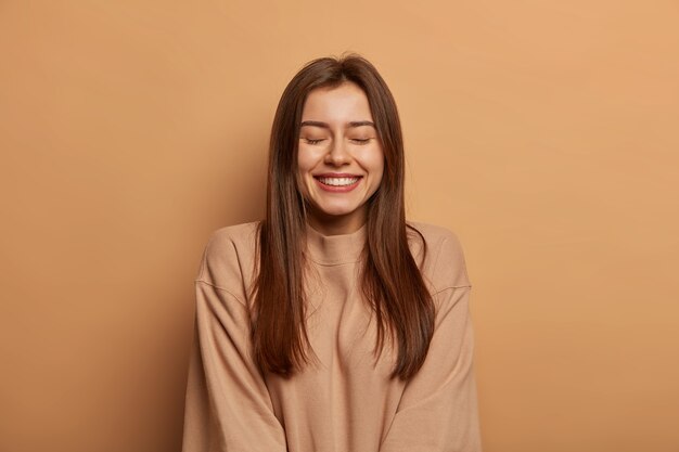 Retrato de mujer hermosa con cabello lacio oscuro, sonríe ampliamente, mantiene los ojos cerrados, disfruta de un momento agradable