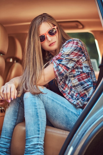 Foto gratuita retrato de una mujer hermosa con una camisa de lana y jeans, sentada en el auto en el asiento trasero con una puerta abierta.