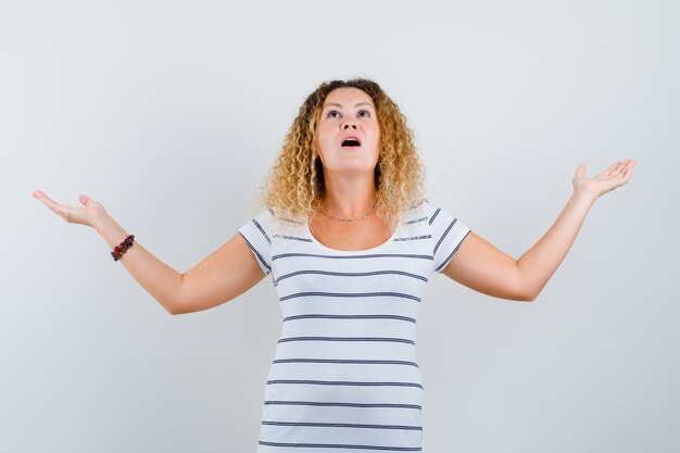 Retrato de mujer hermosa extendiendo las palmas a un lado mientras mira hacia arriba en camiseta y mira asombrada vista frontal