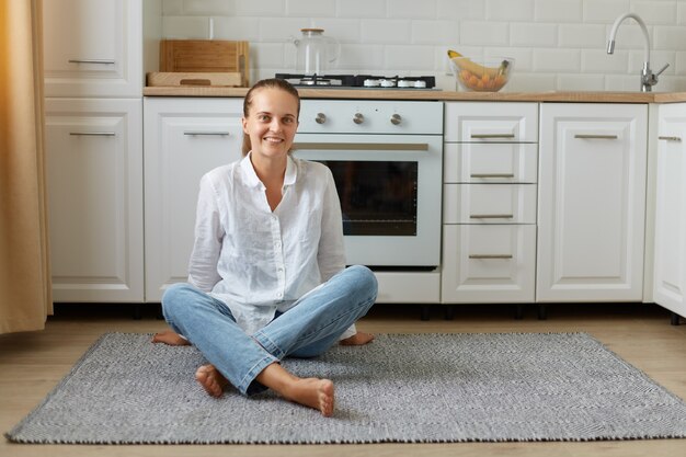 Retrato de mujer hermosa feliz posando interior, mirando a la cámara mientras está sentado en el piso de la cocina en casa, chica con cola de caballo vestida con jeans y camisa blanca.