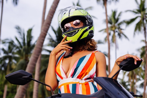 Retrato de mujer hermosa jinete en casco de moto verde amarillo y vestido de verano luz colorida en selva en campo tropical bajo palmeras.