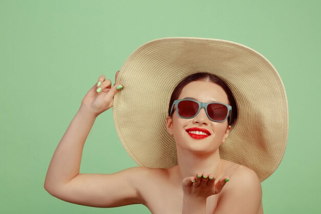 Retrato de mujer hermosa con maquillaje brillante, gafas rojas y sombrero en espacio verde. Maquillaje elegante y de moda, peinado. Belleza, moda y concepto publicitario