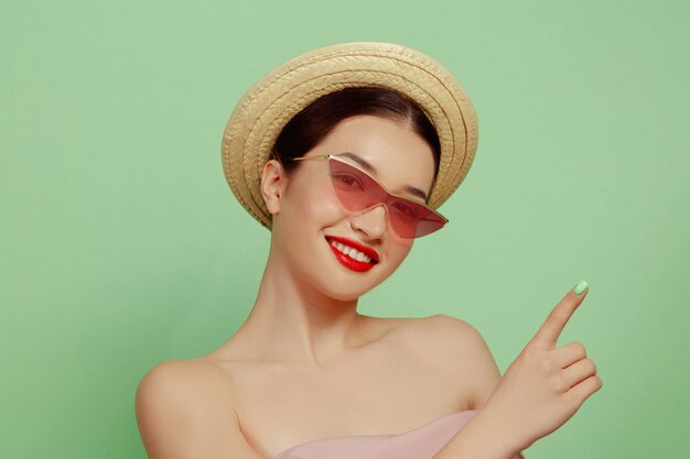 Retrato de mujer hermosa con maquillaje brillante, gafas rojas y sombrero en estudio verde