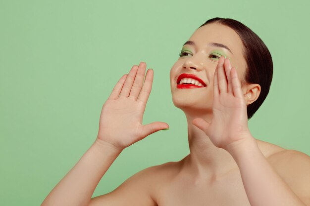 Retrato de mujer hermosa con maquillaje brillante, gafas rojas y sombrero sobre fondo verde de estudio. Maquillaje elegante y de moda, peinado. Concepto de belleza, moda y publicidad. Llamar a ventas, sonriendo.