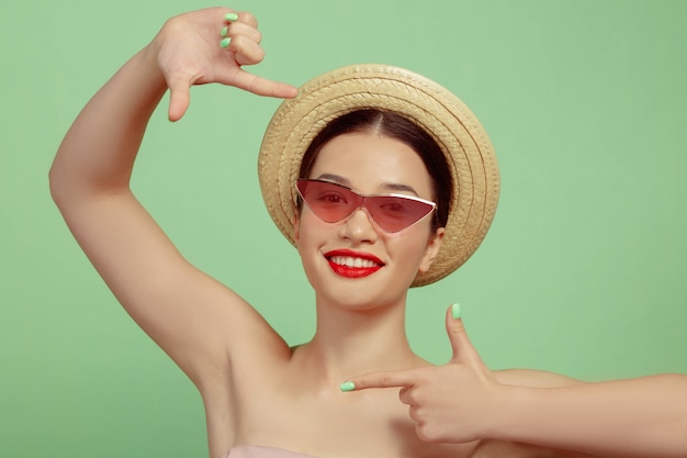 Retrato de mujer hermosa con maquillaje brillante, gafas rojas y sombrero sobre fondo verde de estudio. Maquillaje elegante y de moda, peinado. Concepto de belleza, moda y publicidad. Sonriendo, haciendo un tiro.