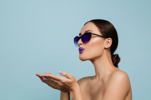 Retrato de mujer hermosa con maquillaje brillante y gafas de sol sobre fondo azul de estudio. Maquillaje y peinado con estilo y de moda. Colores de verano. Concepto de belleza, moda y publicidad. Mandando besos.