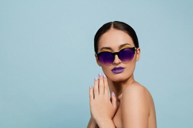 Retrato de mujer hermosa con maquillaje brillante y gafas de sol sobre fondo azul de estudio. Maquillaje y peinado con estilo y de moda. Colores de verano. Concepto de belleza, moda y publicidad. Serio, confiado.