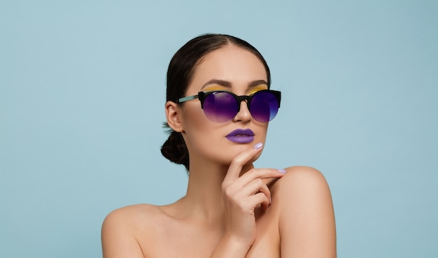 Retrato de mujer hermosa con maquillaje brillante y gafas de sol sobre fondo azul de estudio. Maquillaje y peinado con estilo y de moda. Colores de verano. Concepto de belleza, moda y publicidad. Serio, confiado.